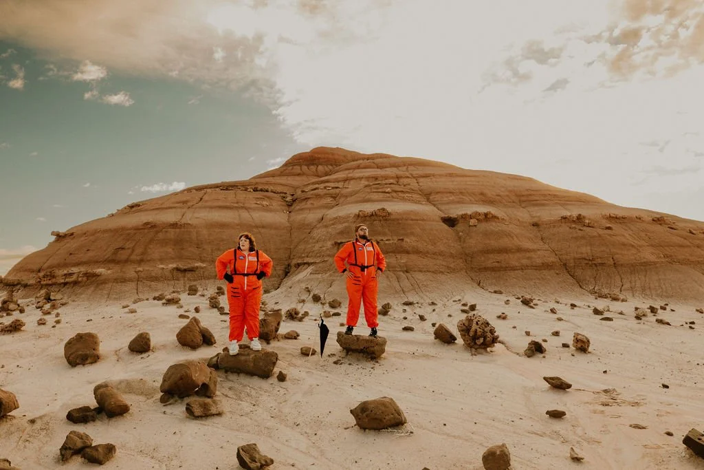 A newlywed couple dressed as astronauts in orange space suits standing on rocks in the Utah desert.