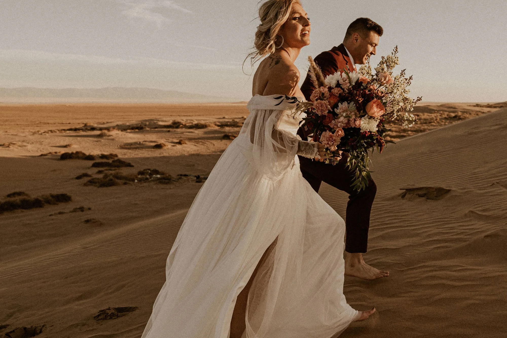 A bride and groom walking barefoot in a desert at sunset. The bride is wearing a white off-shoulder wedding dress and holding a large bouquet of flowers. The groom is dressed in dark pants and a suit jacket.