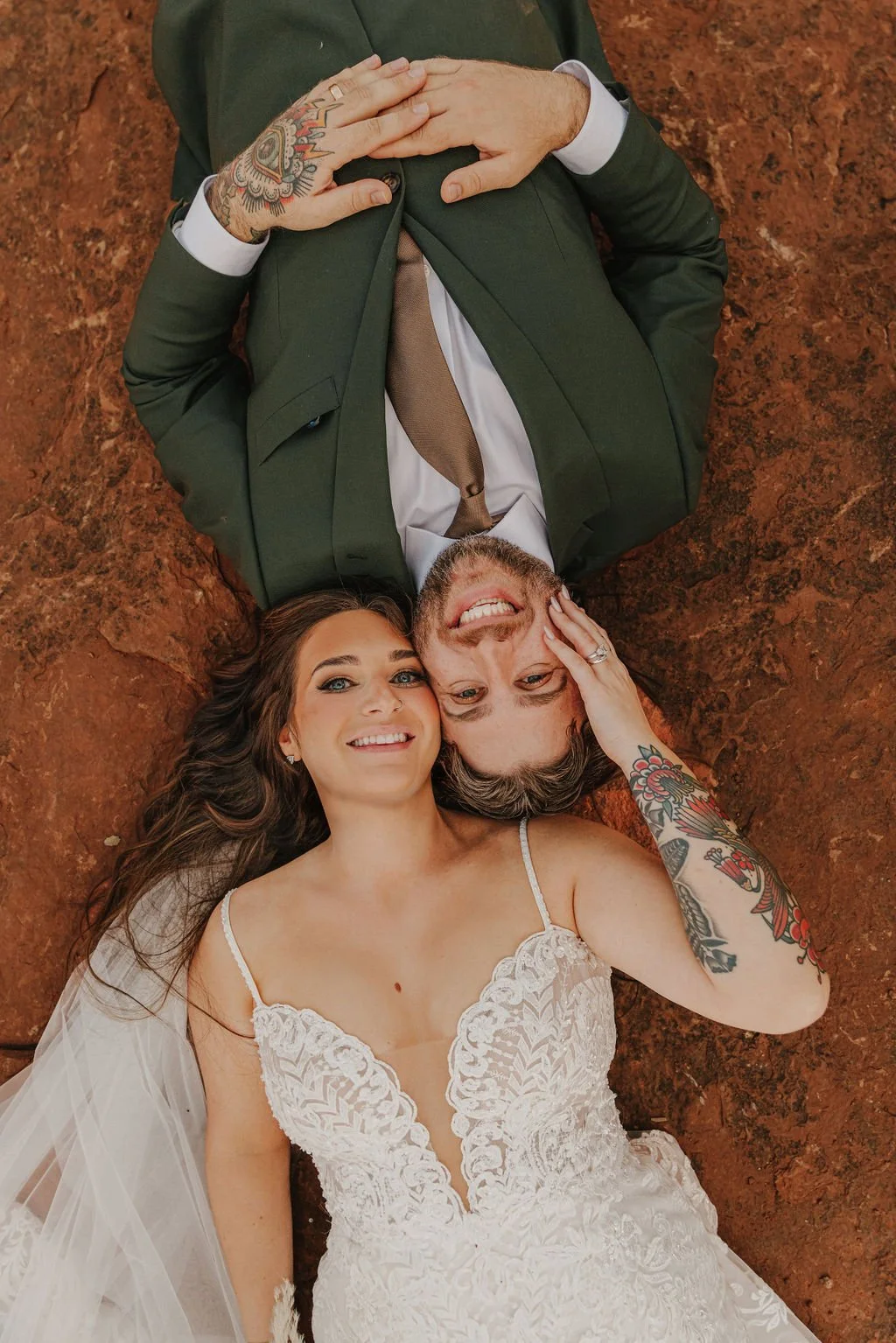 A man and woman lying on red rock, smiling and looking up at the camera, with his head resting on her arm. The woman is wearing a white lace wedding dress, and the man is dressed in a green suit with a tie. 