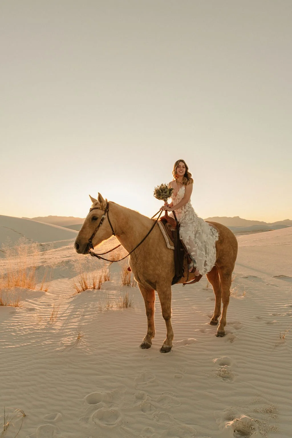 A woman in a white lace wedding dress riding a tan horse through a desert landscape at sunset, holding a bouquet of flowers.