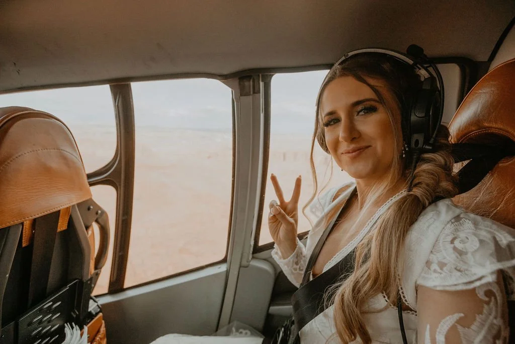 Young woman with long hair, wearing a white lace dress and headphones, smiling and making a peace sign inside a helicopter with desert landscape outside.