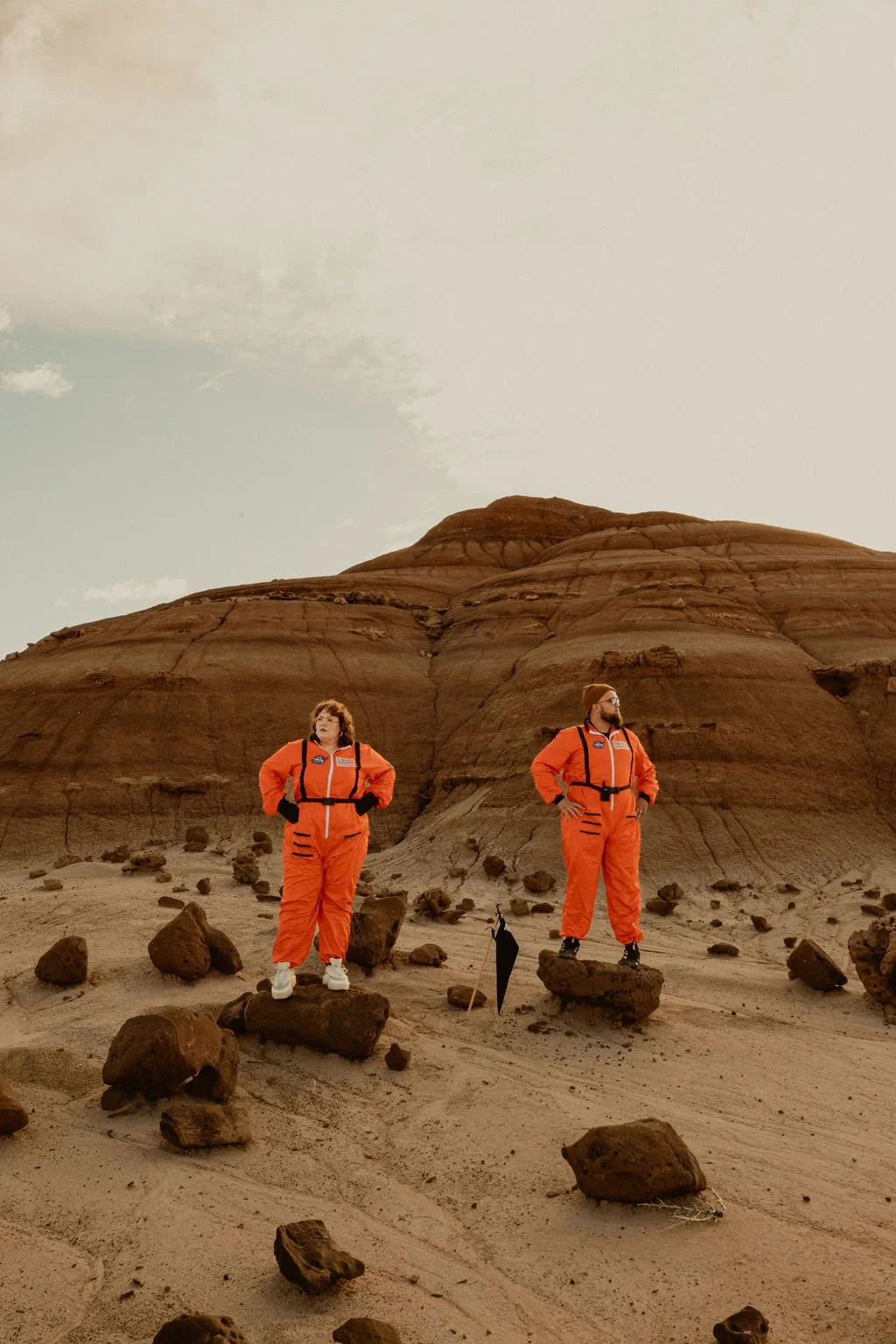 Two people in orange space suits standing on rocks in a desert landscape with a large rock formation in the background.