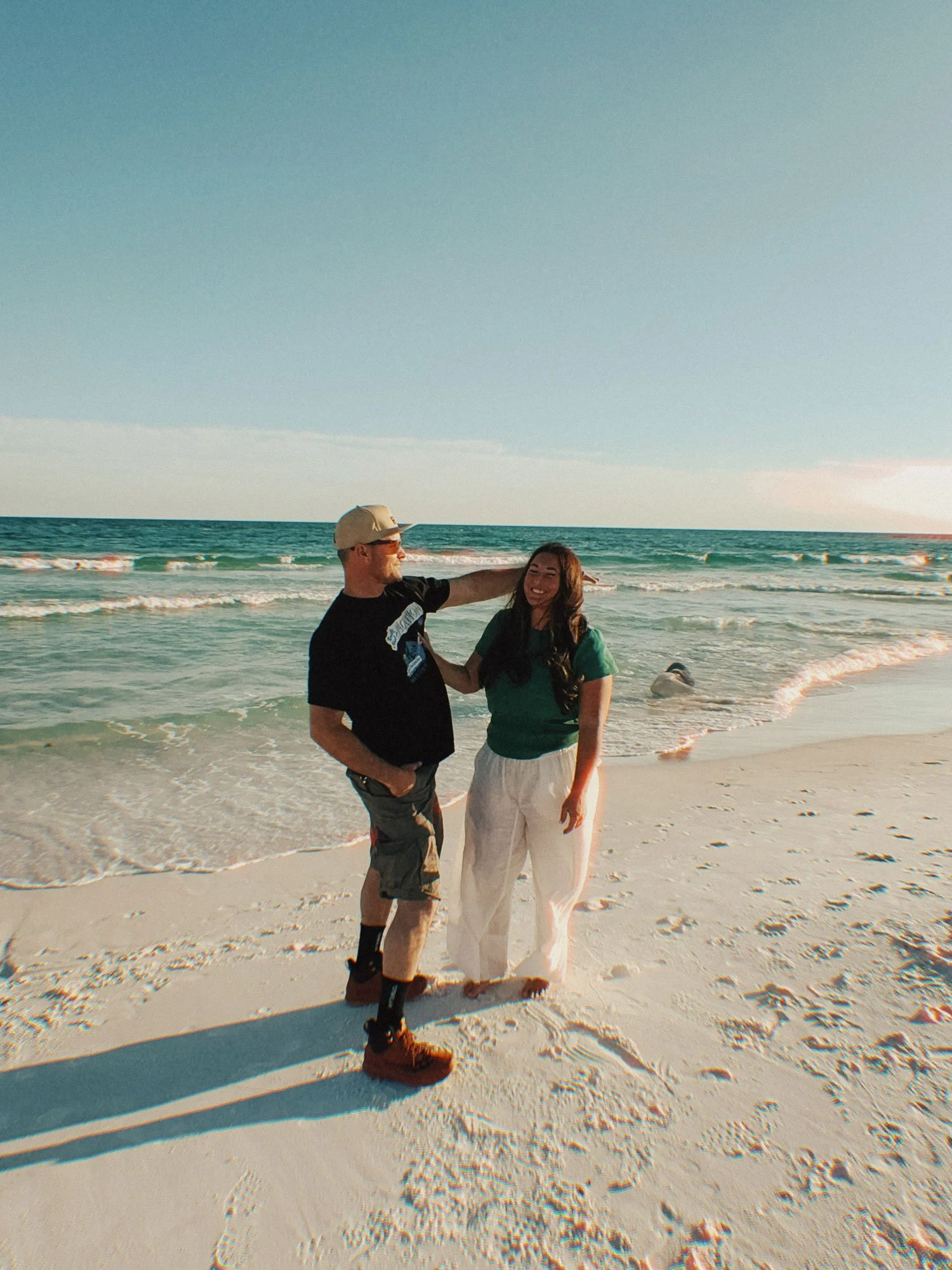 A man and a woman stand on a sandy beach near the water, smiling and talking, with the ocean and a clear sky in the background.