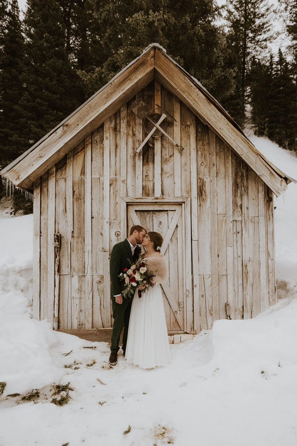 A bride and groom standing close together in front of a rustic wooden shed in a snowy outdoor setting, sharing a tender moment. The bride holds a large bouquet of flowers, and both are dressed in wedding attire.