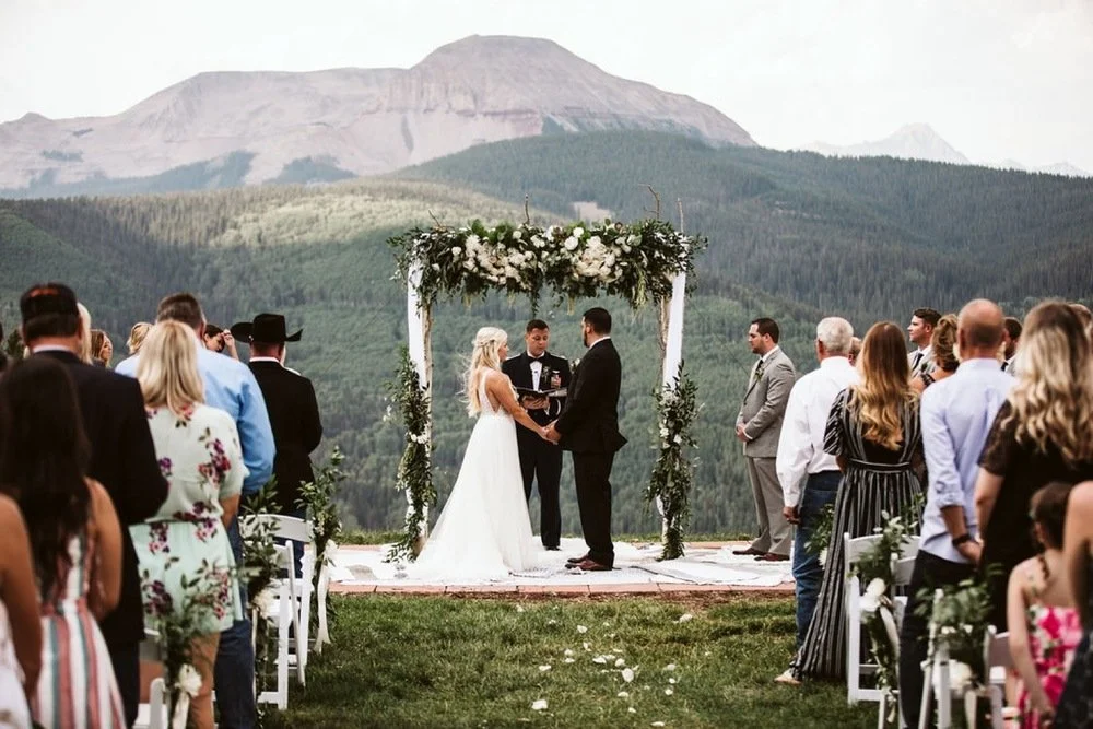 Couple getting married under a floral arch at an outdoor wedding ceremony, with guests standing on either side, Colorado mountains in the background.