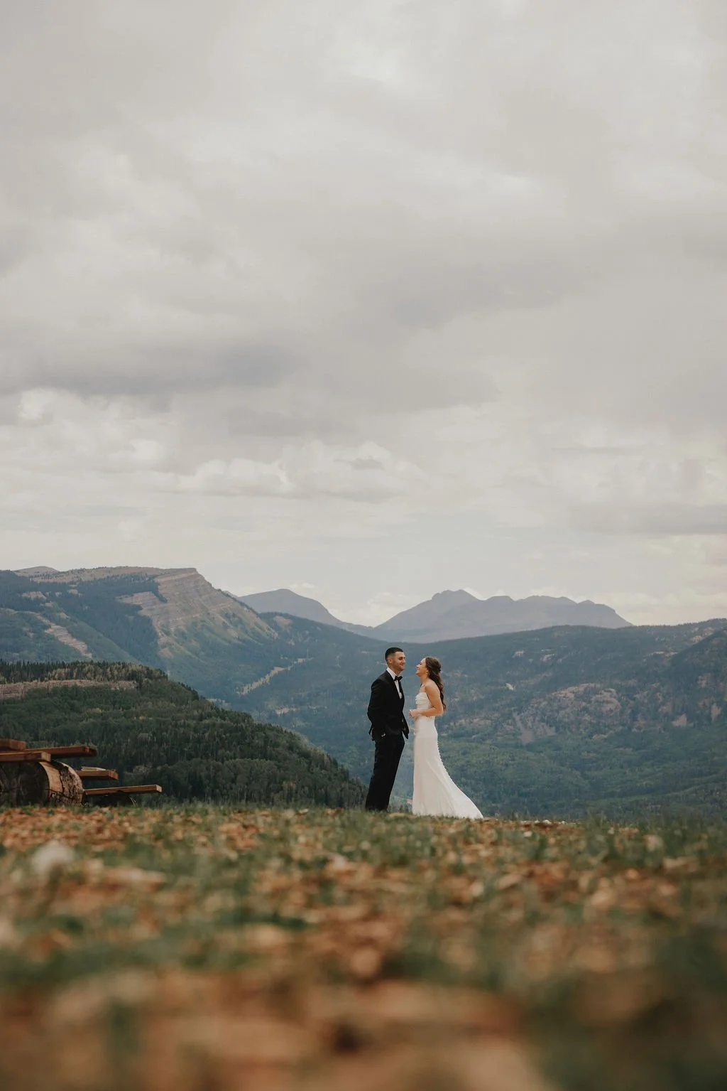 A bride and groom in wedding attire standing on a grassy hill with mountains in the background. Overcast sky.
