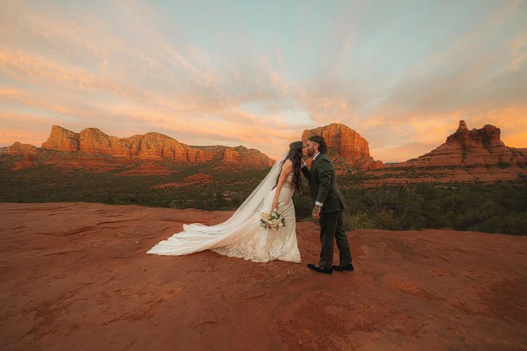 A bride and groom in wedding attire kissing on a red rock formation at sunset with desert mountains in the background.