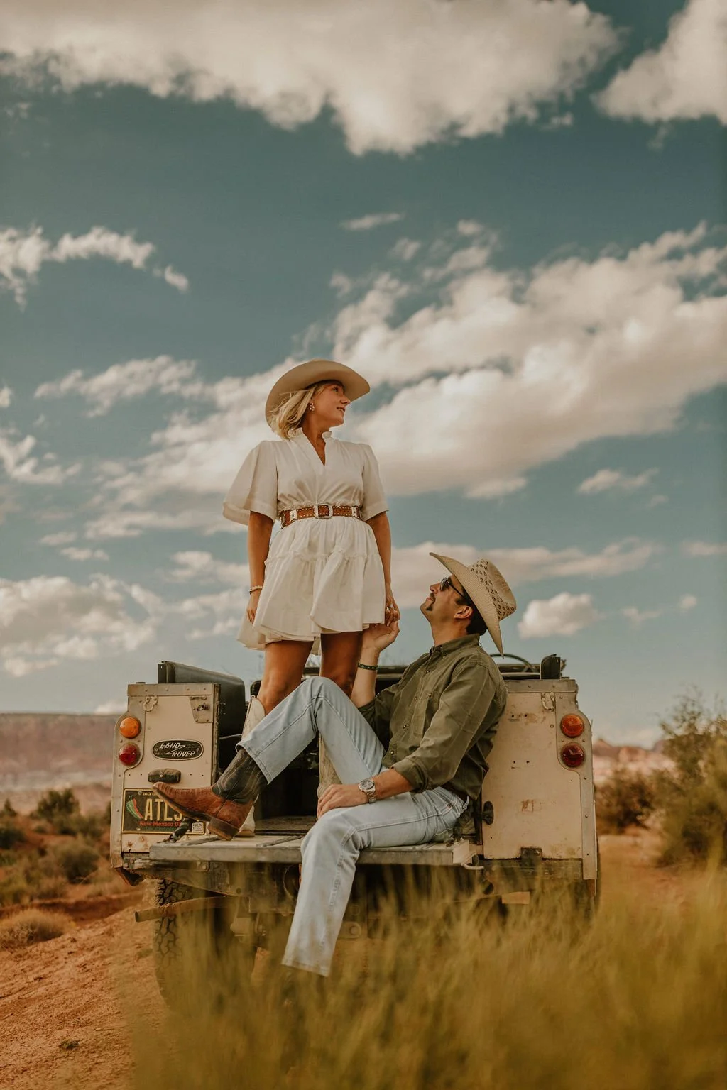 A couple dressed in Western attire sitting on the back of a vintage Land Rover in a desert landscape with a blue sky and white clouds.
