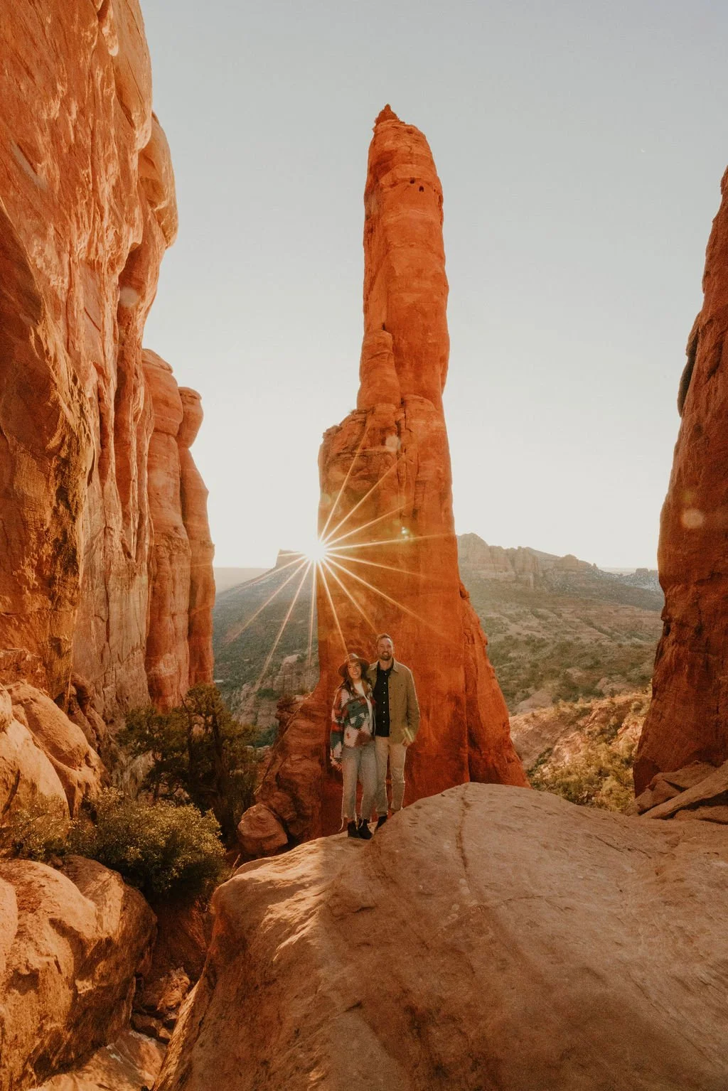 Two people standing on a large rock in a desert canyon with tall red rock formations. The sun is setting behind a prominent towering spire, creating a starburst effect.