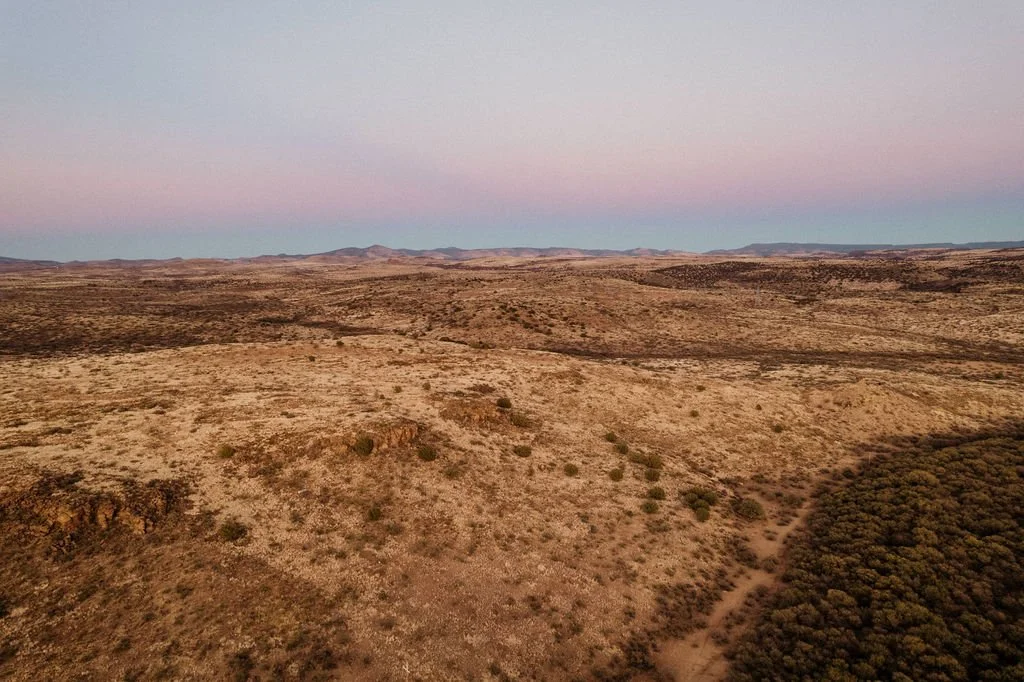 A vast desert landscape at sunset with rolling hills, sparse vegetation, and a colorful sky with pink and blue hues.