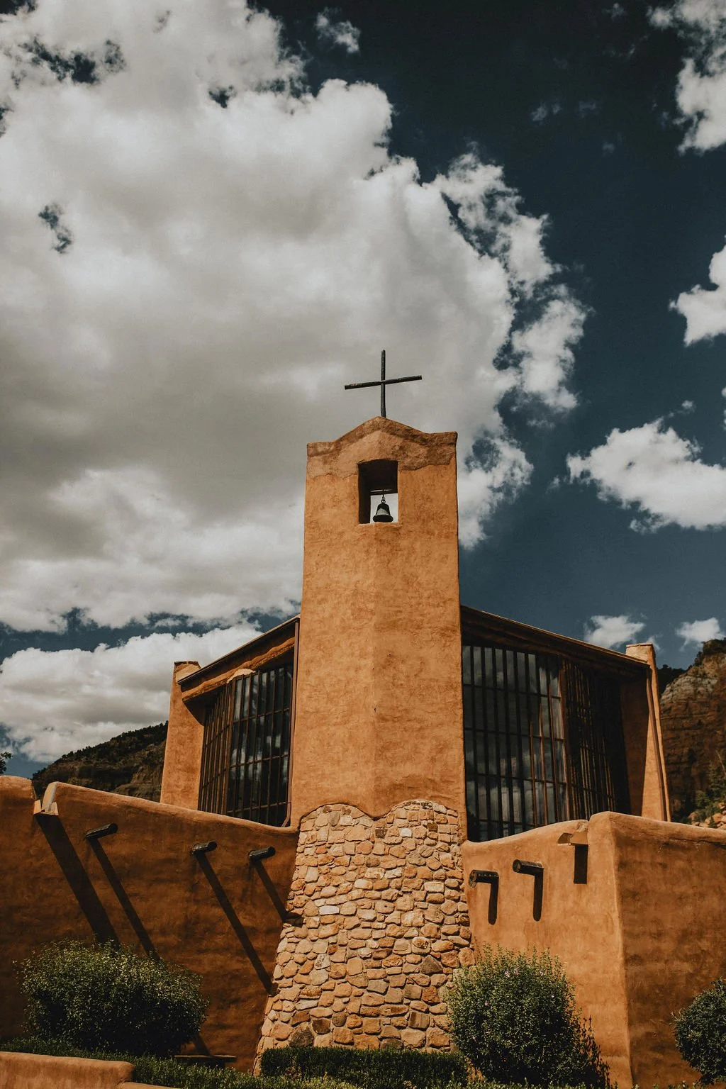 A church with a bell tower topped by a cross, against a blue sky with clouds, surrounded by desert landscape and small bushes.