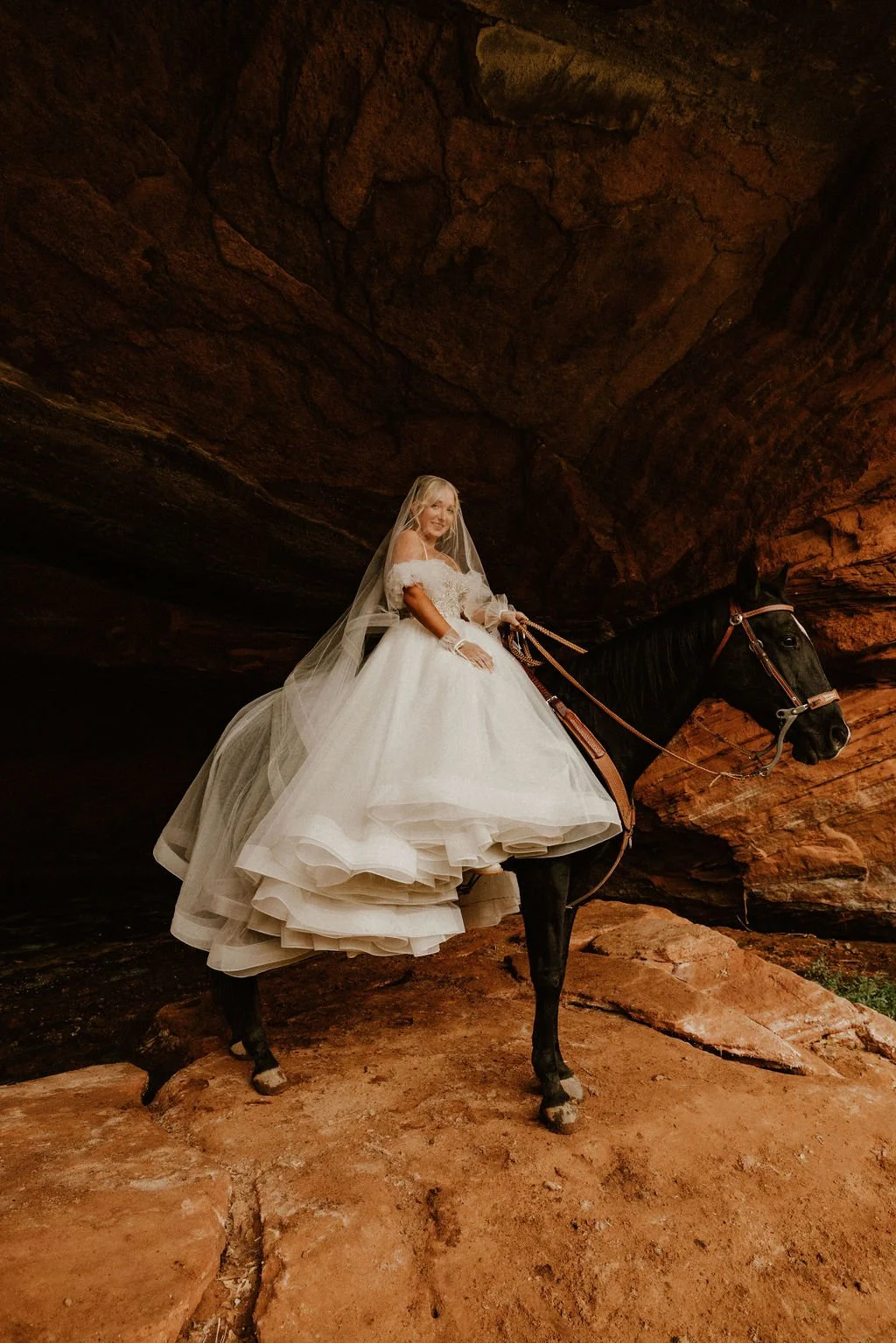 A bride in a white wedding gown and veil riding a black horse inside a rocky cave.