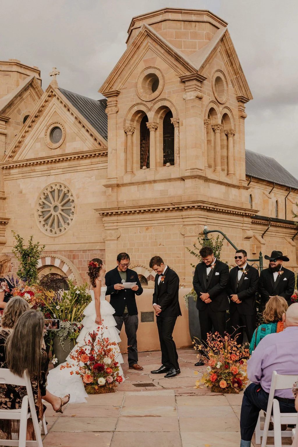 A wedding ceremony taking place outdoors in front of a historic stone church with a large rose window and bell tower. The bride and groom stand with officiant and groomsmen, surrounded by floral arrangements and seated guests.
