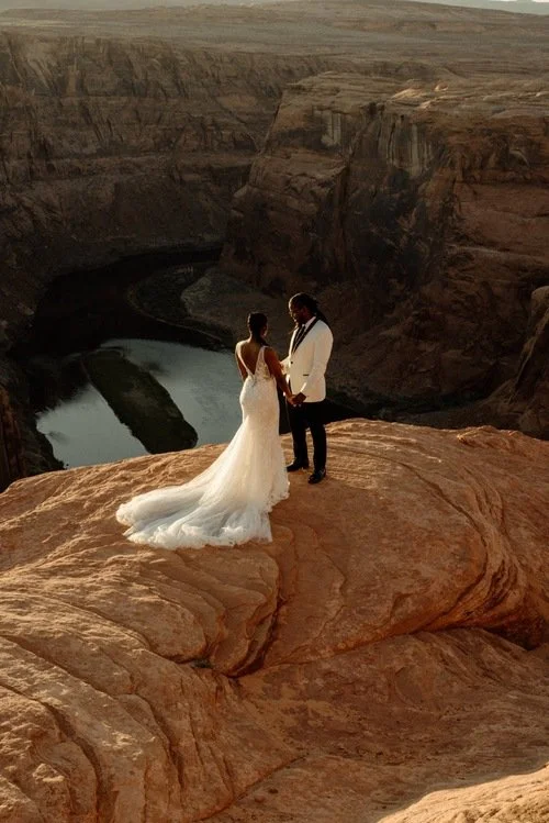 A bride and groom holding hands on a rocky cliff overlooking a river canyon at sunset.