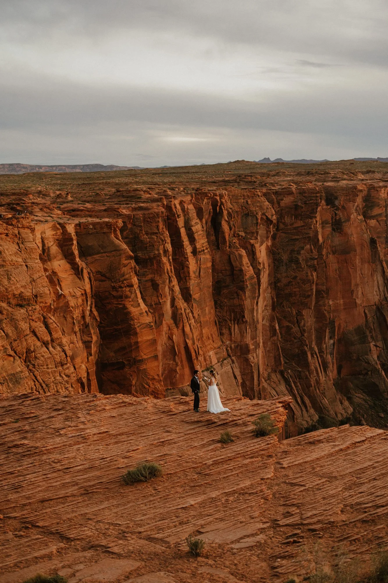 A couple in wedding attire standing on a rocky cliff edge overlooking a deep canyon with reddish rock formations.