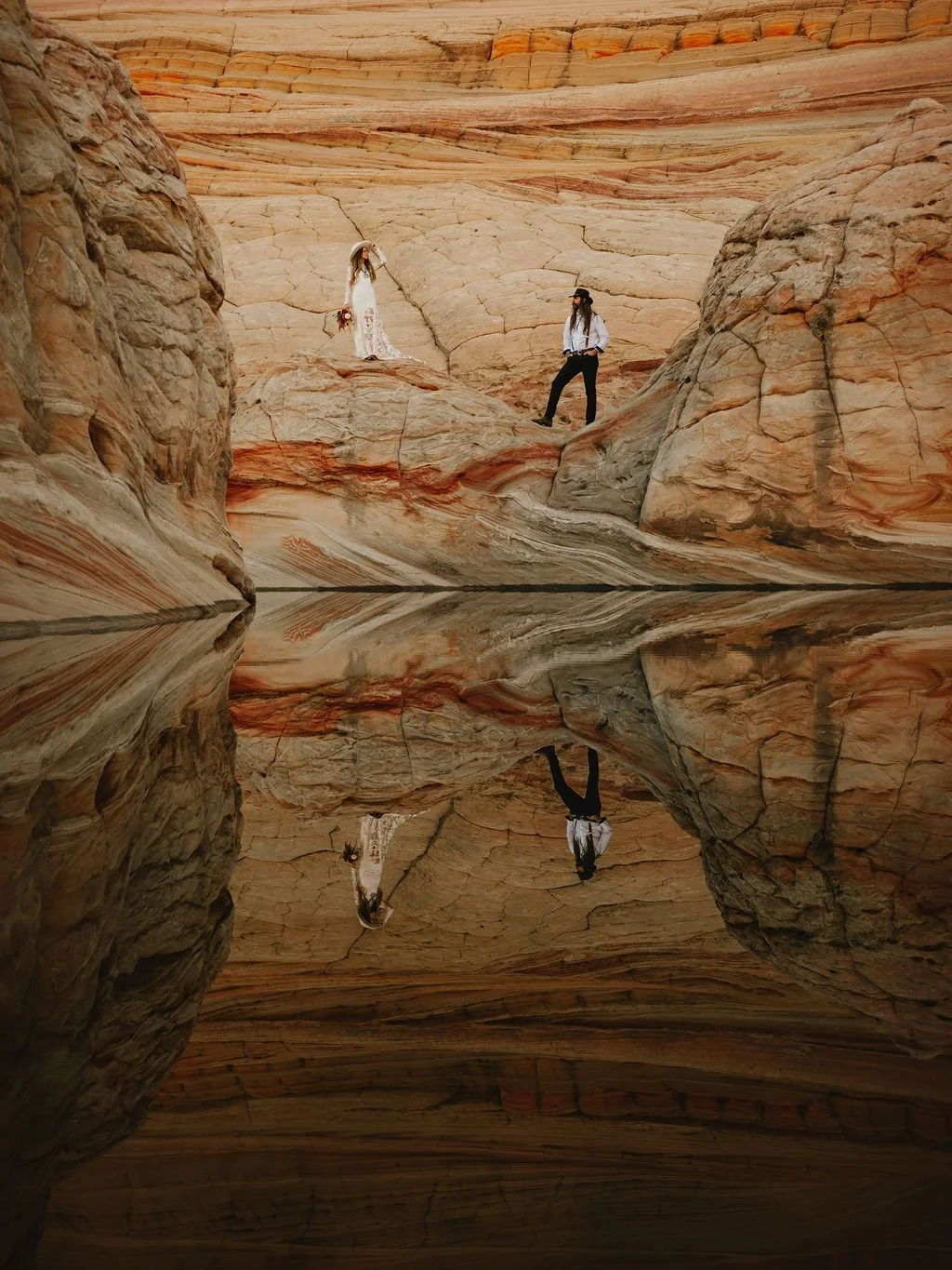 A couple standing on rocks inside a canyon with striped sandstone walls, their reflection visible in a calm body of water at the bottom of the image.