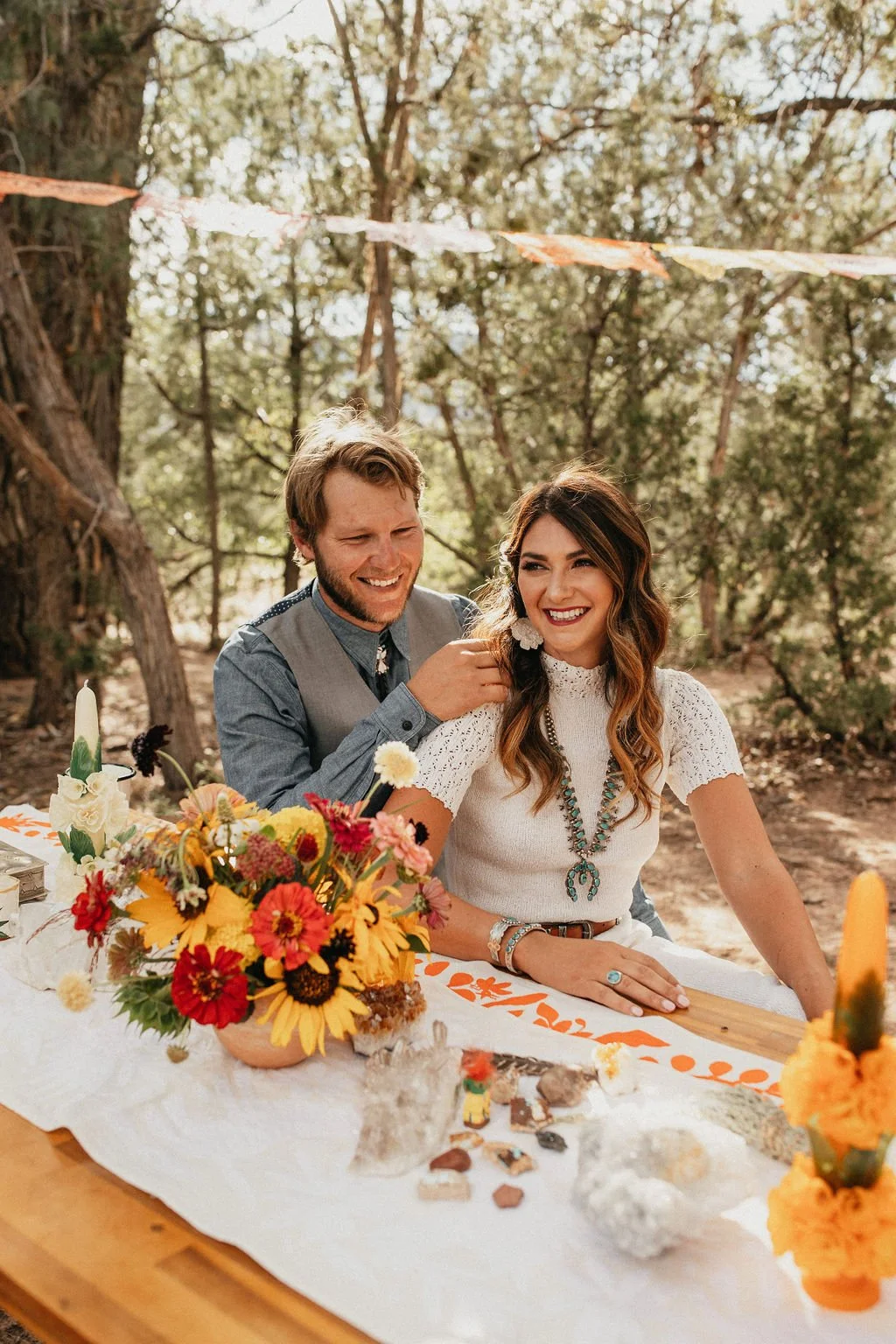 A couple at an outdoor wedding reception, smiling and sitting at a table decorated with a floral centerpiece, surrounded by trees and string lights.