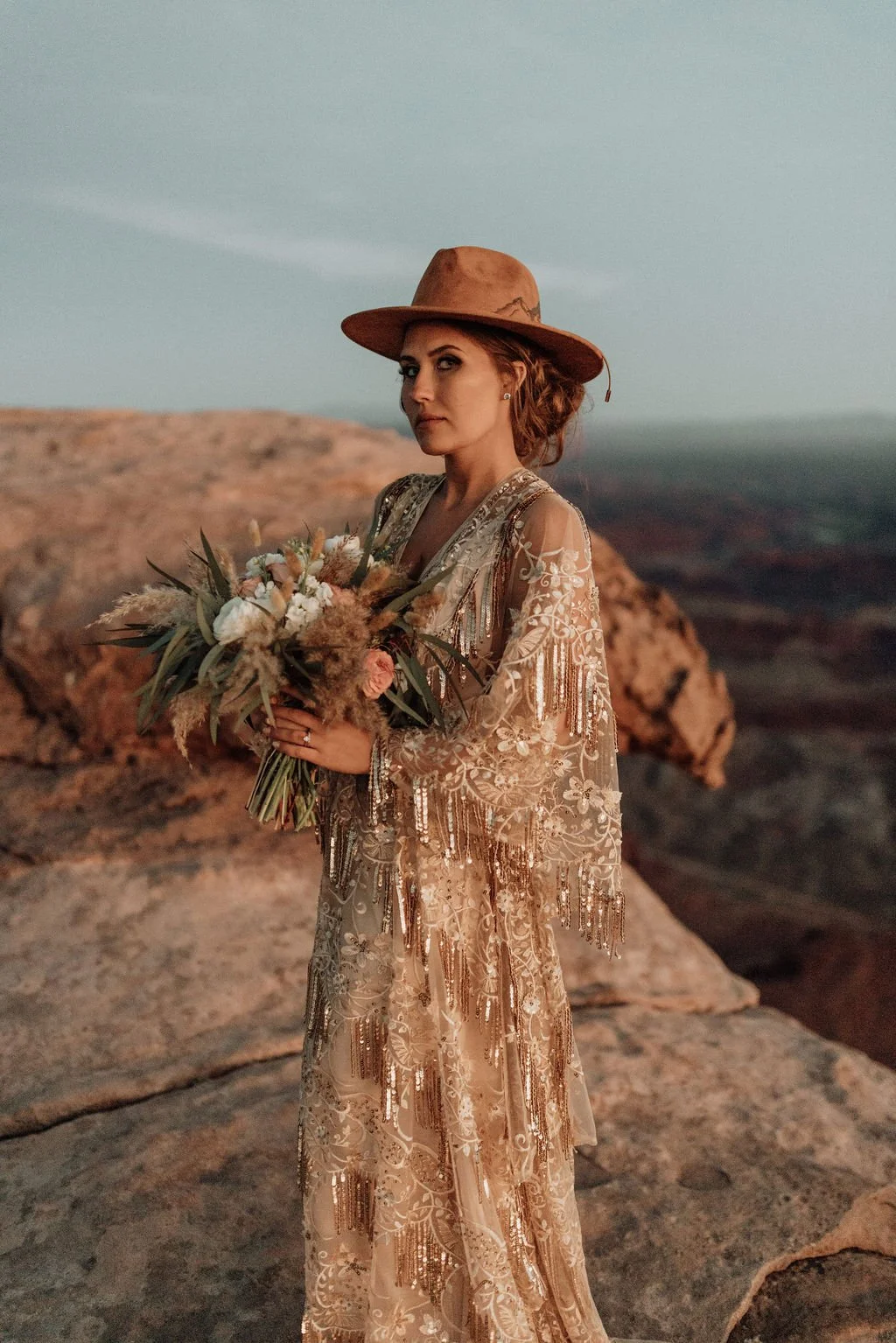 A woman in a long, ornate, embroidered dress and wide-brimmed hat holds a bouquet of flowers while standing on rocky terrain during sunset or twilight.