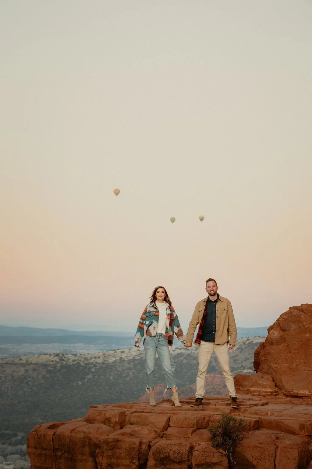 A couple standing hand in hand on a rocky cliff at sunset, with hot air balloons floating in the sky above them.