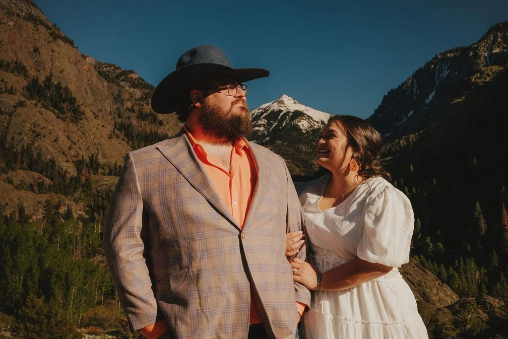 A man with a beard and glasses wearing a large black cowboy hat, a plaid blazer, and an orange shirt, stands next to a woman in a white dress, both outdoors in a mountainous landscape with snowy peaks and trees.