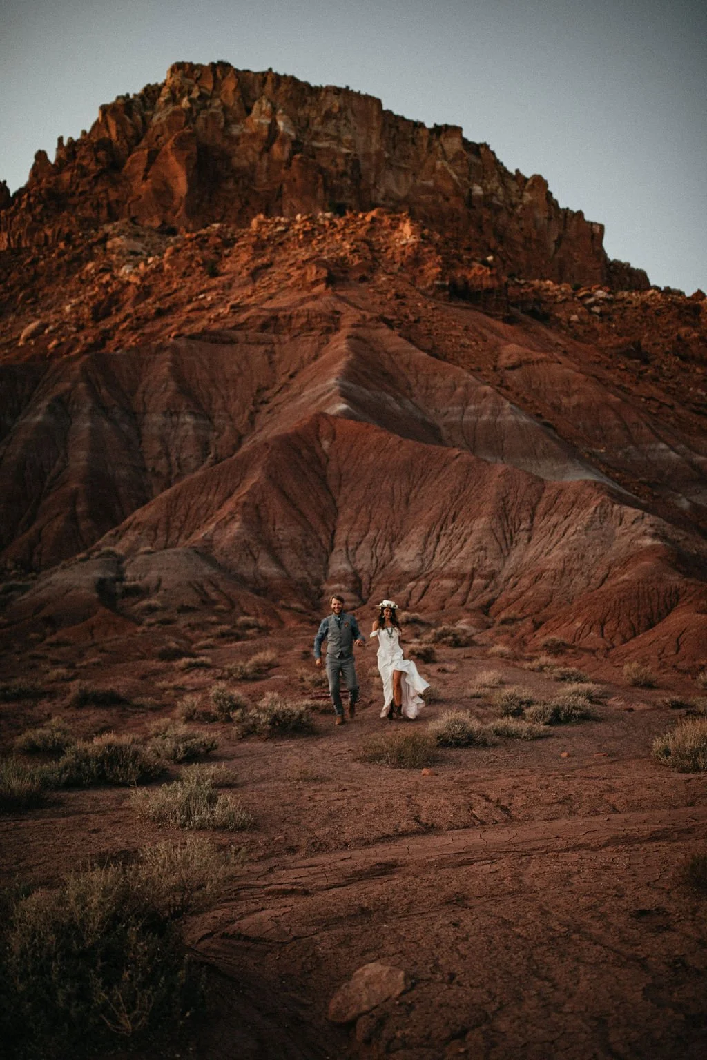 A couple dressed in wedding attire walking hand in hand through a desert landscape with rugged, reddish mountains in the background at sunset or dawn.