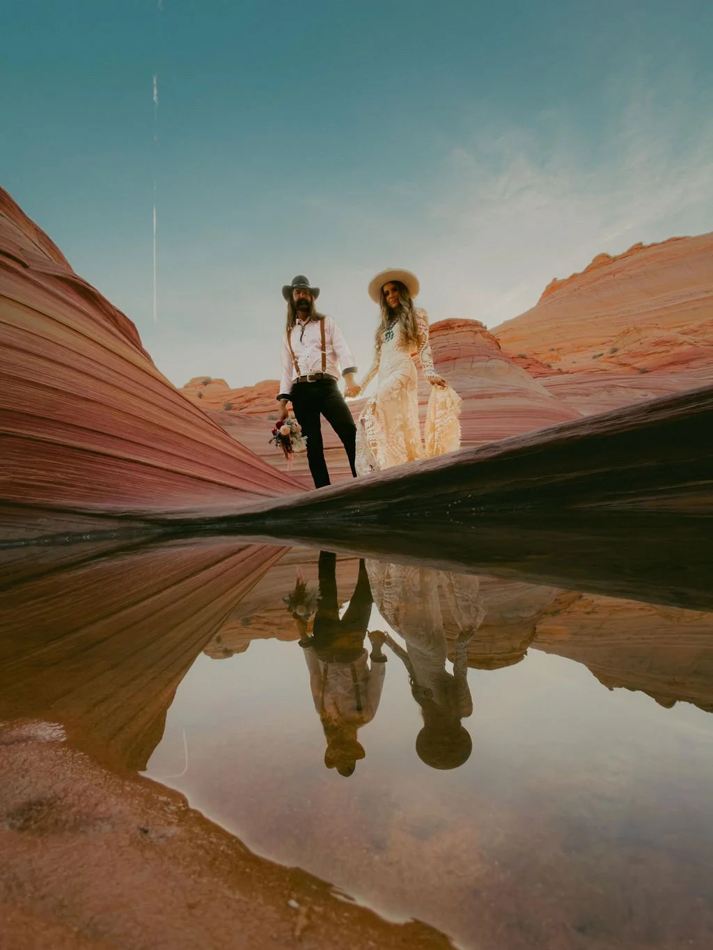 A couple standing on a rock formation in a desert-like landscape with multicolored sandstone formations. The reflection of the couple and rocks can be seen in a small pool of water on the ground. The sky is clear with a faint jet trail visible.