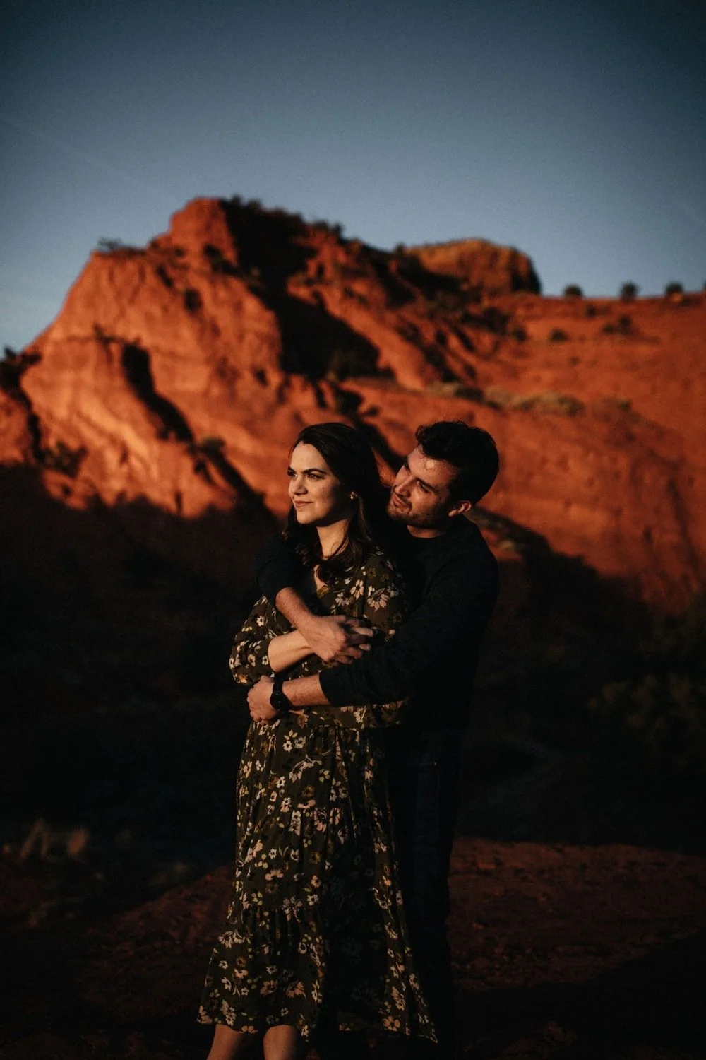 A couple hugging under a sunset sky with rocky, mountainous terrain in the background.