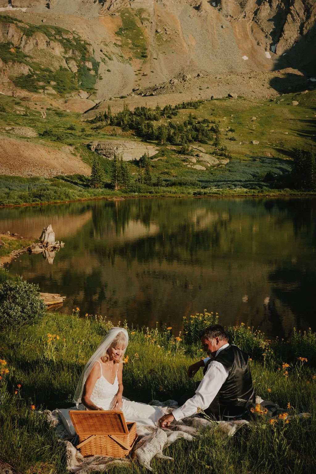 A couple having a picnic by a lake in a mountainous area. The woman is in a white dress with a veil, and the man is in a black vest and white shirt. They are sitting on blankets with a picnic basket, surrounded by yellow flowers, with mountains and a