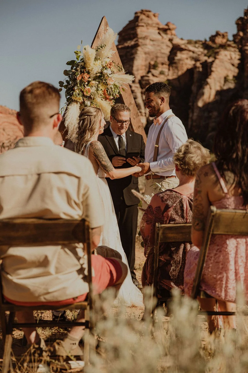 A wedding ceremony outdoors in a desert with rock formations in the background, featuring a bride and groom holding hands and exchanging vows, an officiant, and seated guests.