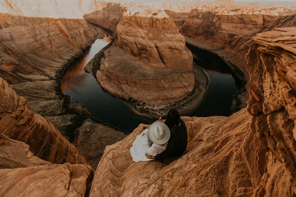 A couple sitting on a rock ledge overlooking a winding river in a canyon at sunset.
