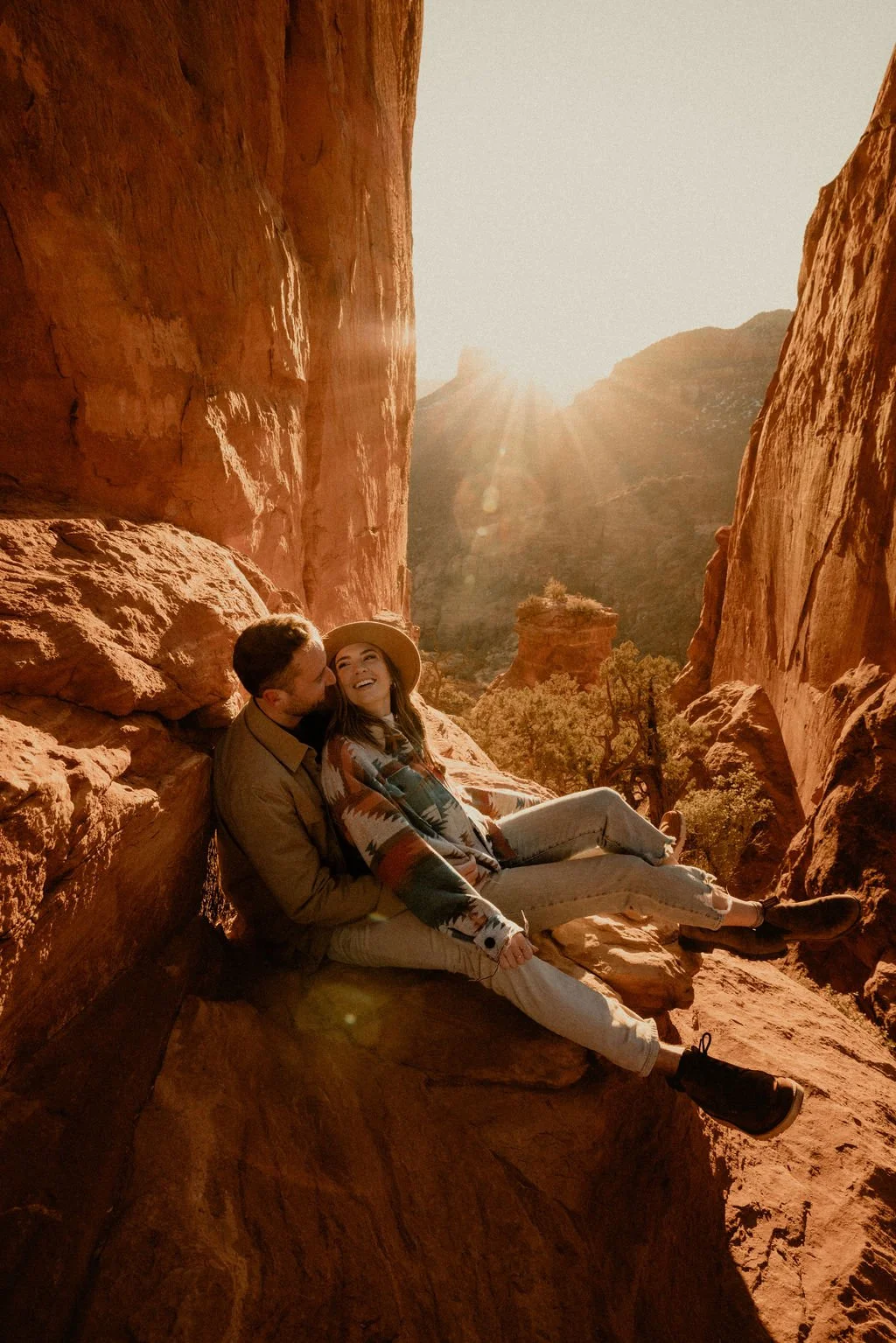 A couple sitting on a large rock in a canyon, with red rock cliffs and a bright sun behind mountains in the distance.