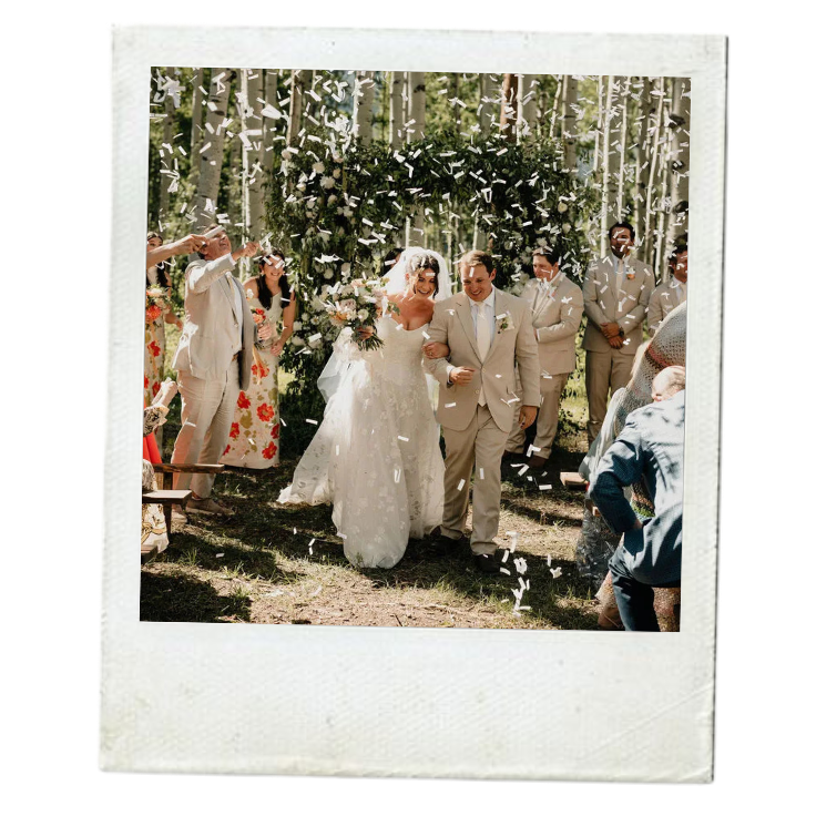 A bride and groom walking down the aisle outdoors, surrounded by celebrating wedding guests and falling confetti, in a forest setting.