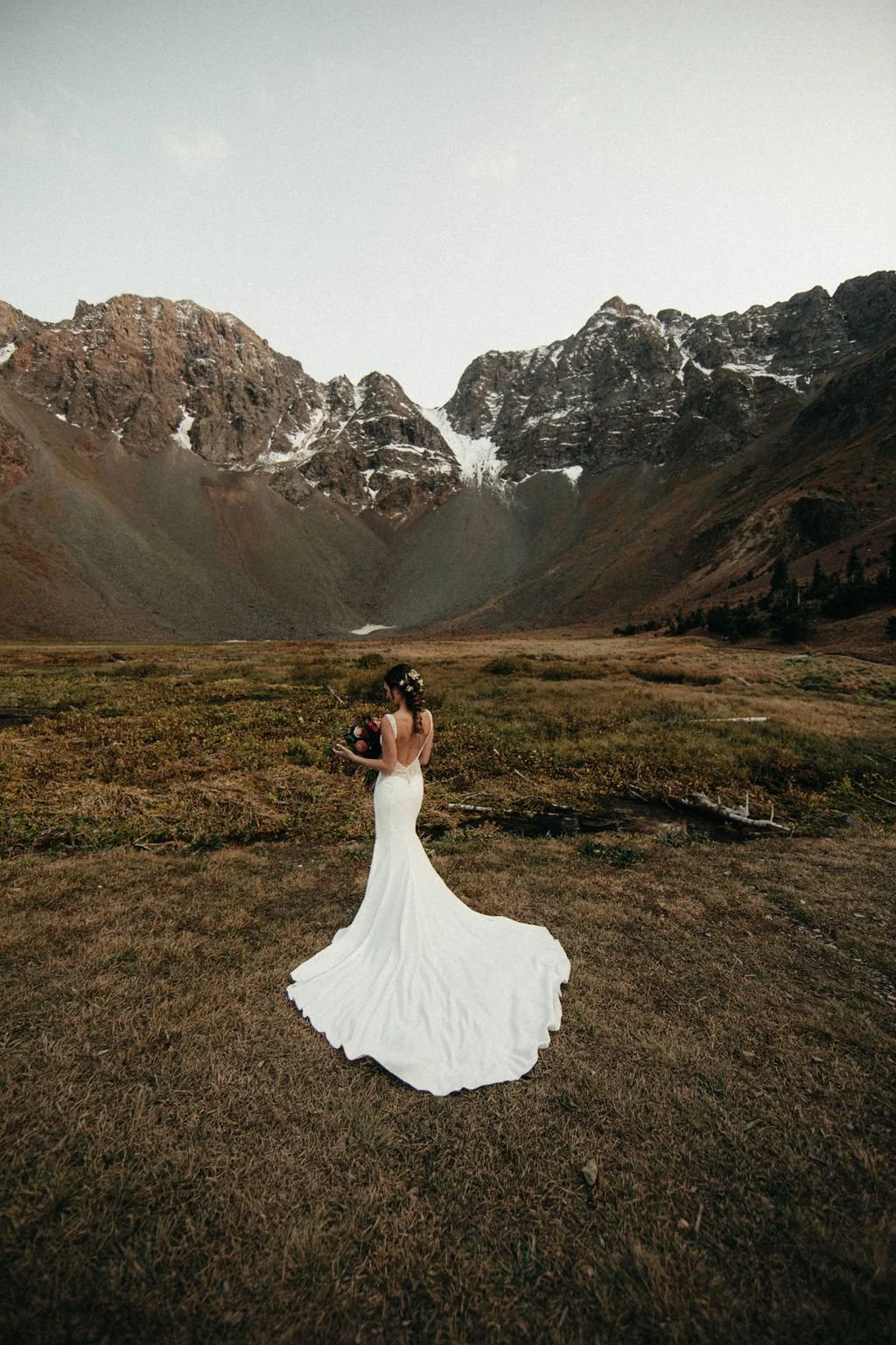 A woman in a white wedding gown holding a bouquet, standing on grassy terrain with wildflowers, with towering mountains in the background and a cloudy sky overhead.