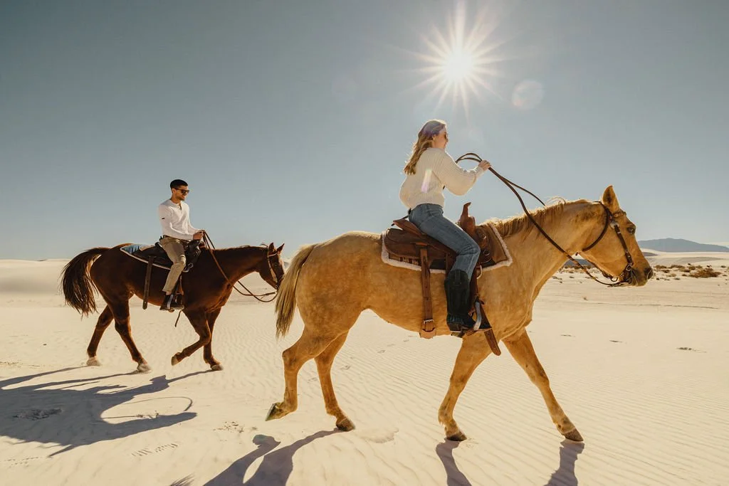 Two people riding horses across a desert landscape with clear skies and a bright sun.
