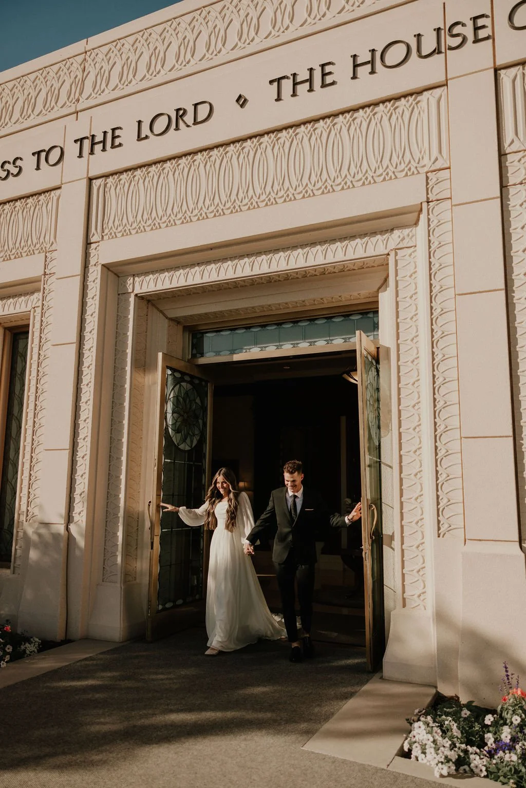 A bride and groom holding hands and walking out of a church with intricate architectural details, stained glass windows, and flowers at the entrance.