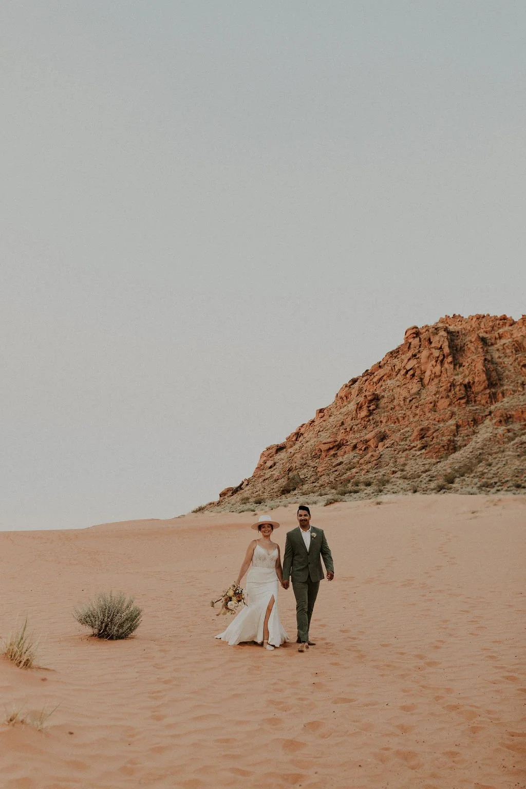 A bride and groom walking hand in hand across a desert landscape with sand dunes and a red rocky hill in the background during daytime.