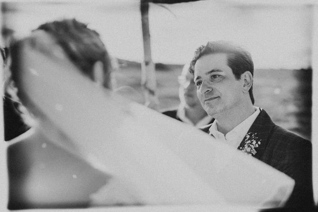 A man with dark hair, dressed in a suit, looking smilingly at a woman under a veil, during a wedding ceremony.