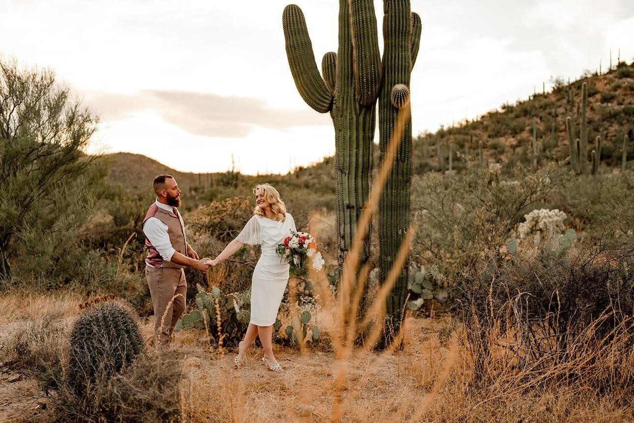 A couple holding hands in a desert landscape with large cacti and dry bushes during sunset, with the woman holding a bouquet of flowers.