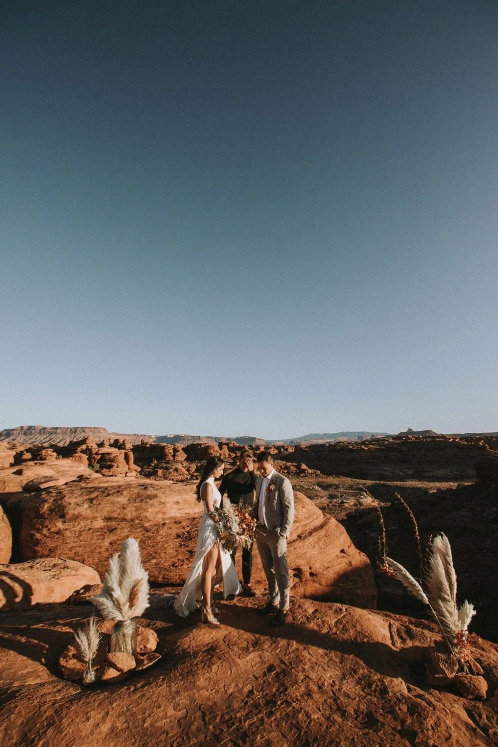 A couple getting married in a desert landscape with rocky formations and pampas grass, under a clear blue sky.