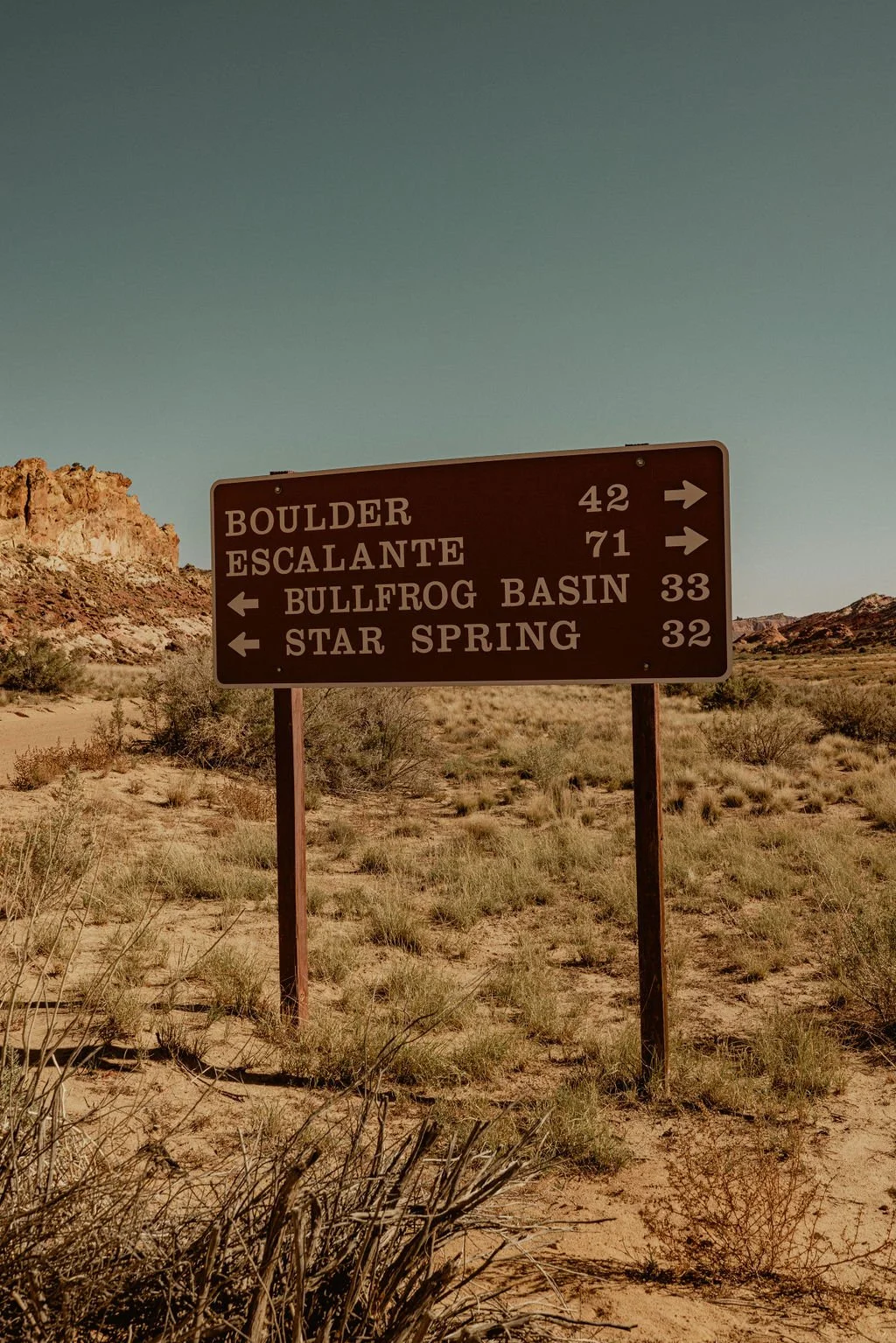 A road sign in a desert landscape pointing to various locations: Boulder (42 miles), Escalante (71 miles) to the right, and Bullfrog Basin (33 miles) and Star Spring (32 miles) to the left.