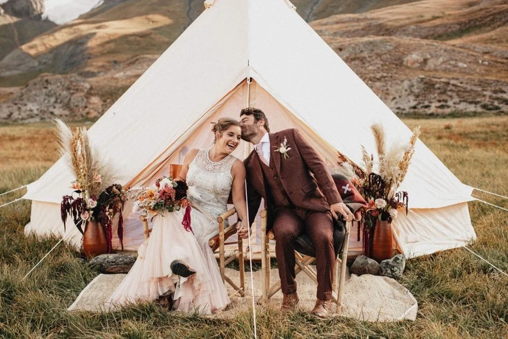 A couple in wedding attire sitting outdoors before a canvas tent, surrounded by decorative vases with flowers and dried plants, in a mountainous landscape.