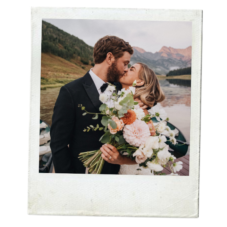 A bride and groom kissing outdoors, with mountains and a lake in the background, during their wedding day.