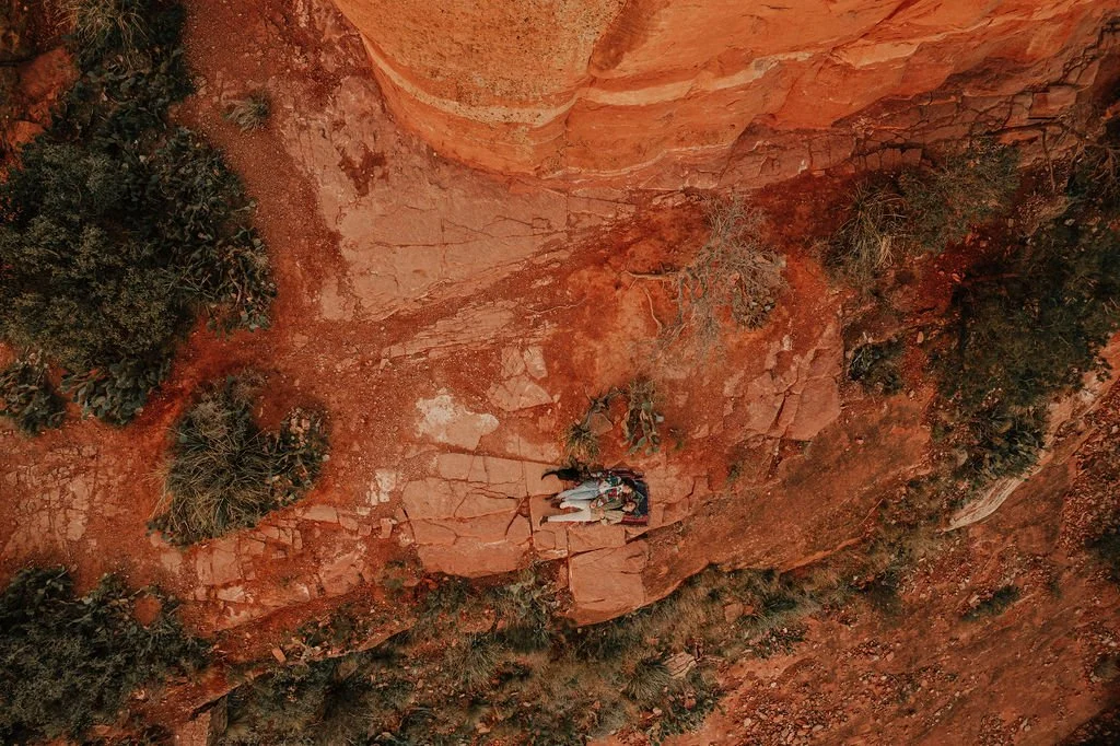 A person climbing rocks on a red desert cliff with sparse green bushes.