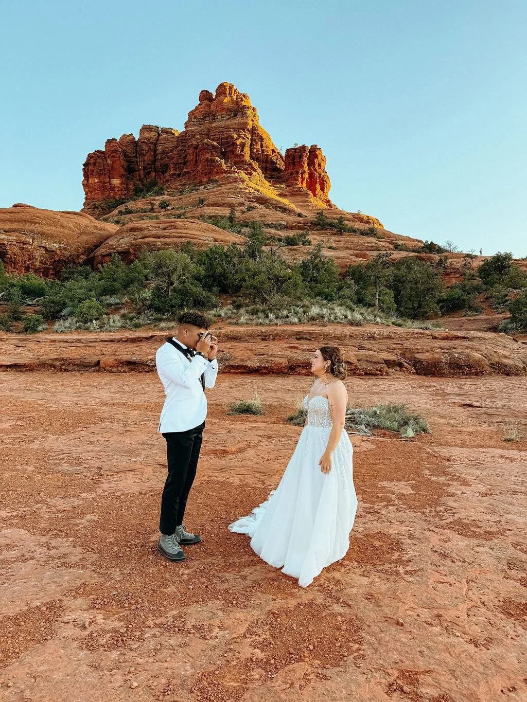 A couple dressed in wedding attire, the bride in a white gown and the groom in a white blazer and black pants, standing on desert terrain with red rocks and a large red rock formation in the background during sunset.