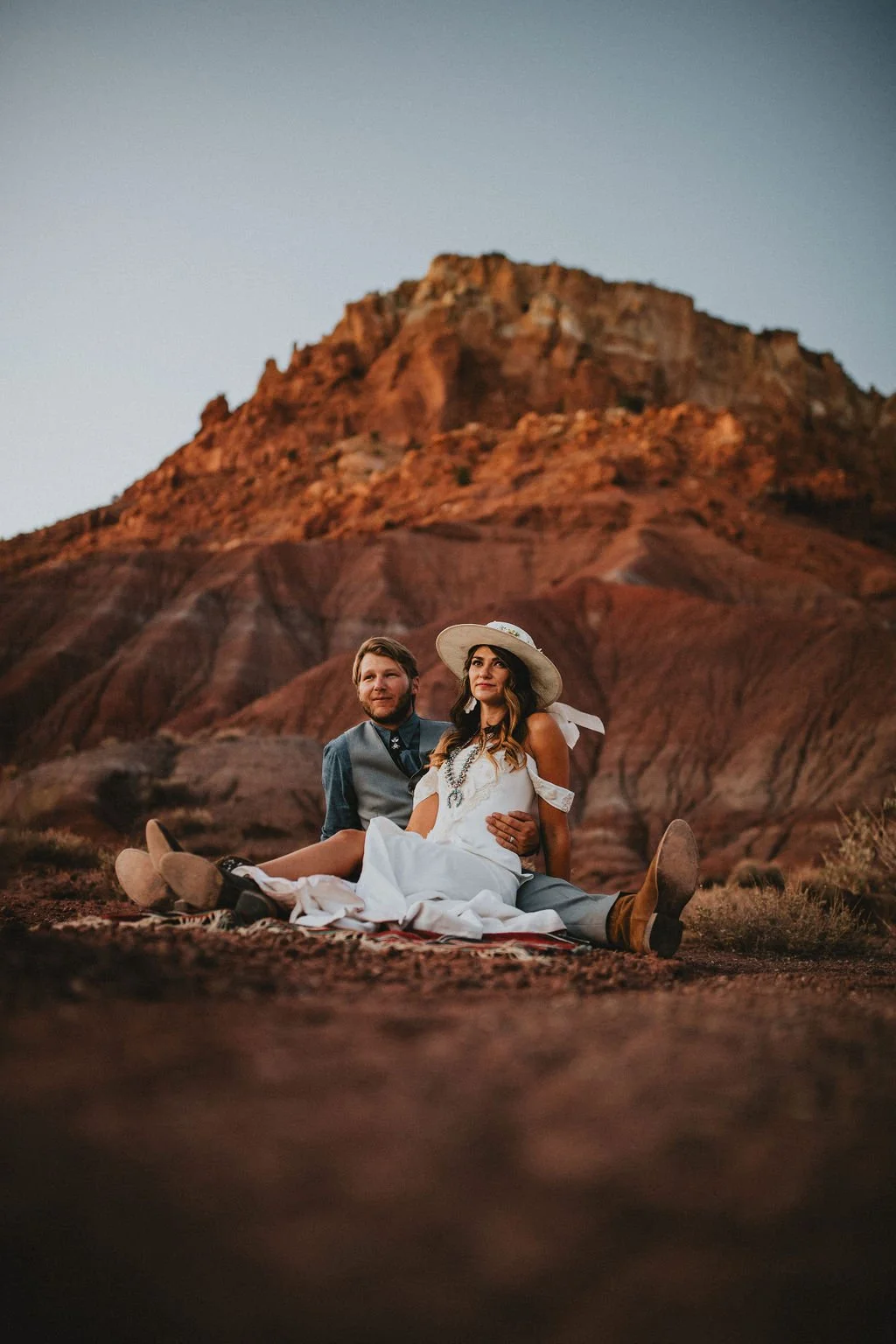 A couple dressed in vintage wedding attire sitting on a blanket on desert ground with a mountainous background at sunset.