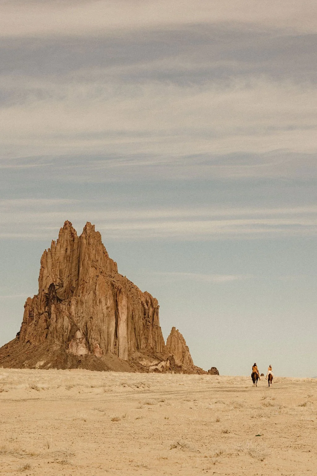 Two people riding horses in a desert landscape in new mexico with a large rocky mountain and a cloudy sky in the background.