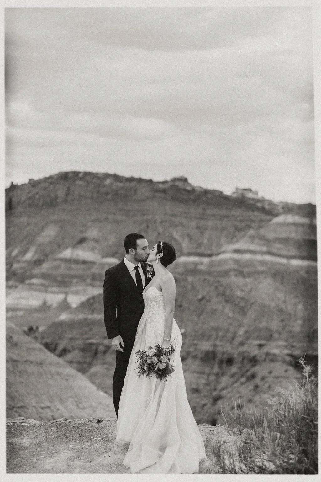 A black and white photo of a newlywed couple kissing in a desert landscape with hills in the background. The bride holds a bouquet of flowers and the groom is dressed in a suit and tie.