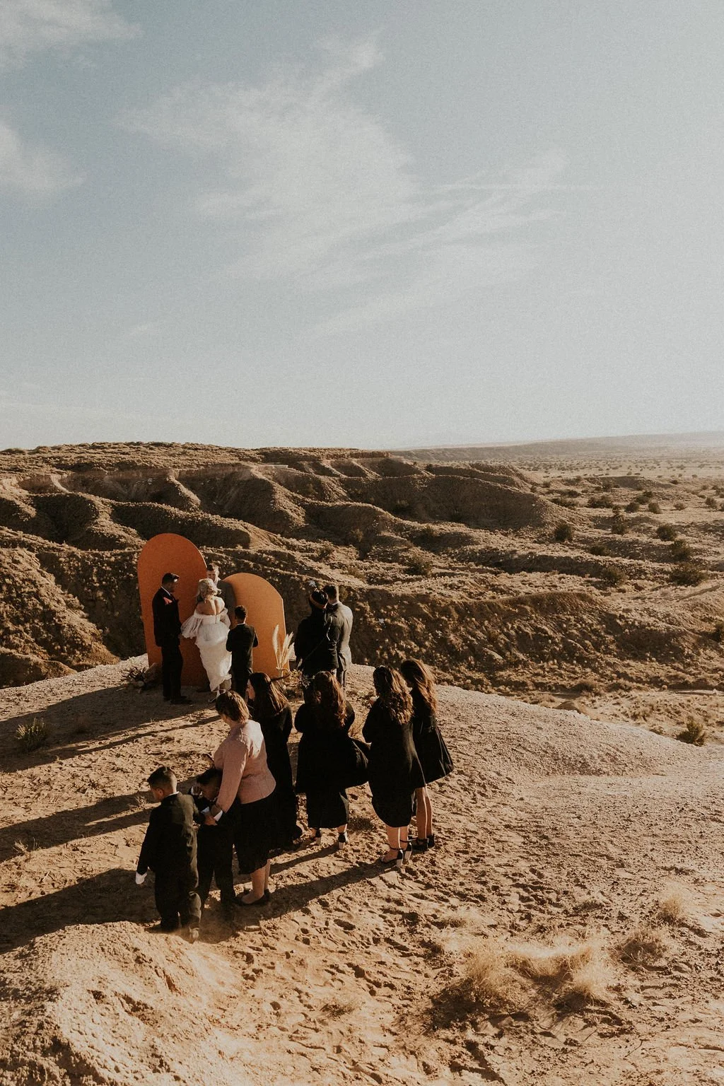 A wedding ceremony taking place in a desert landscape with a small group of guests, an officiant, and a bride and groom standing before large orange panels.