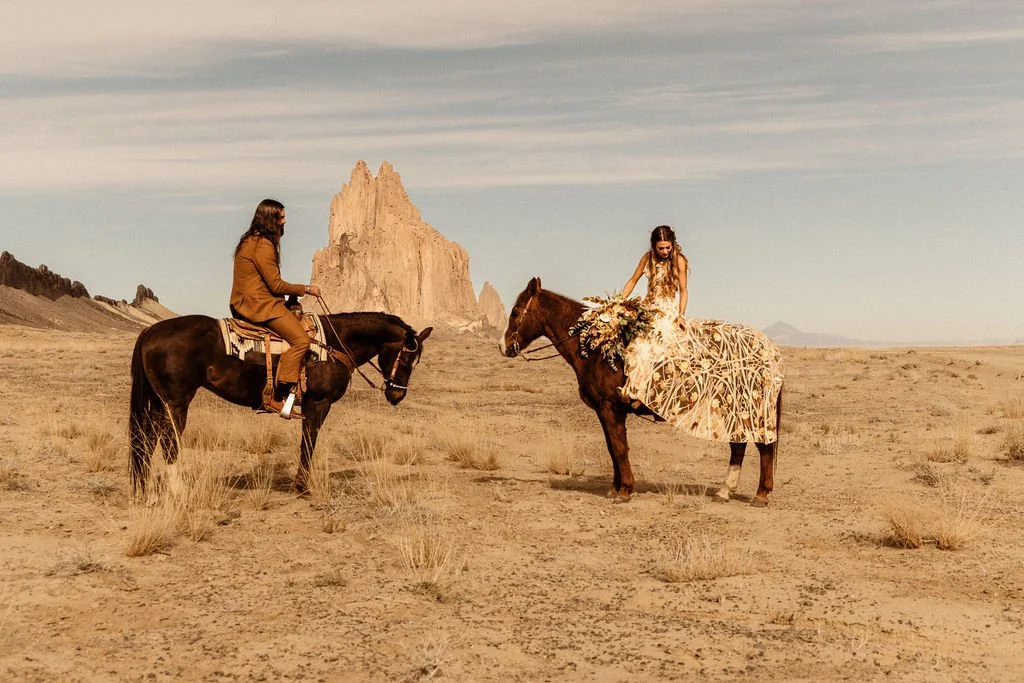 A woman in a floral dress riding a decorated horse, approaching a man in a brown suit riding a black horse in a desert landscape with rock formations in the background in New Mexico.