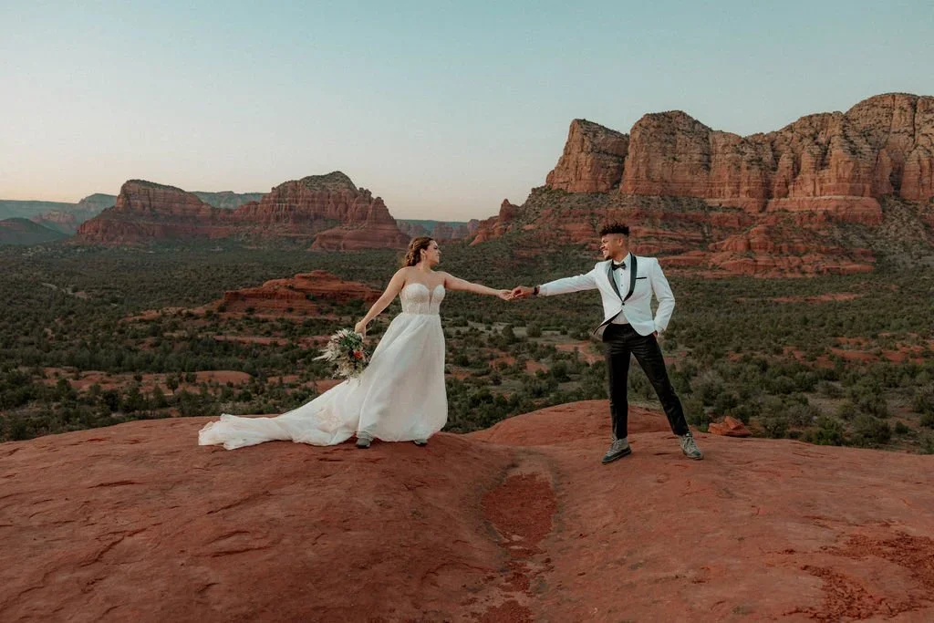 A bride and groom holding hands on a rocky red landscape with red cliffs in the background during sunset.